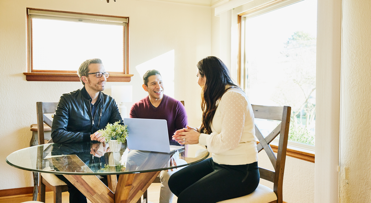 sales person in leather jacket sitting at a glass topped table with a smiling couple looking at a laptop computer