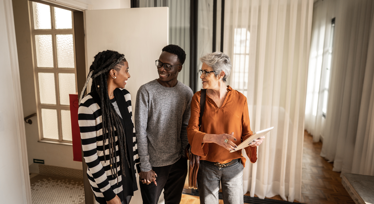 inside a home a millennial couple holding hands and smiling at each other while a middle aged woman stands next to them holding a tablet