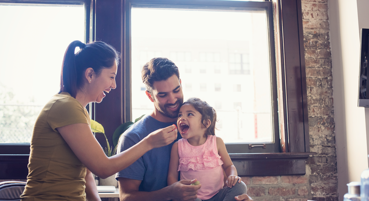 a smiling father holds a little girl while her mother feeds her by hand