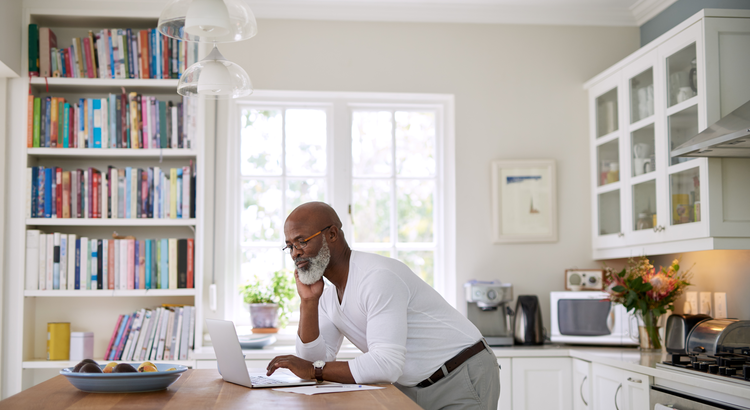 middle aged man standing at a kitchen island looking at a laptop computer